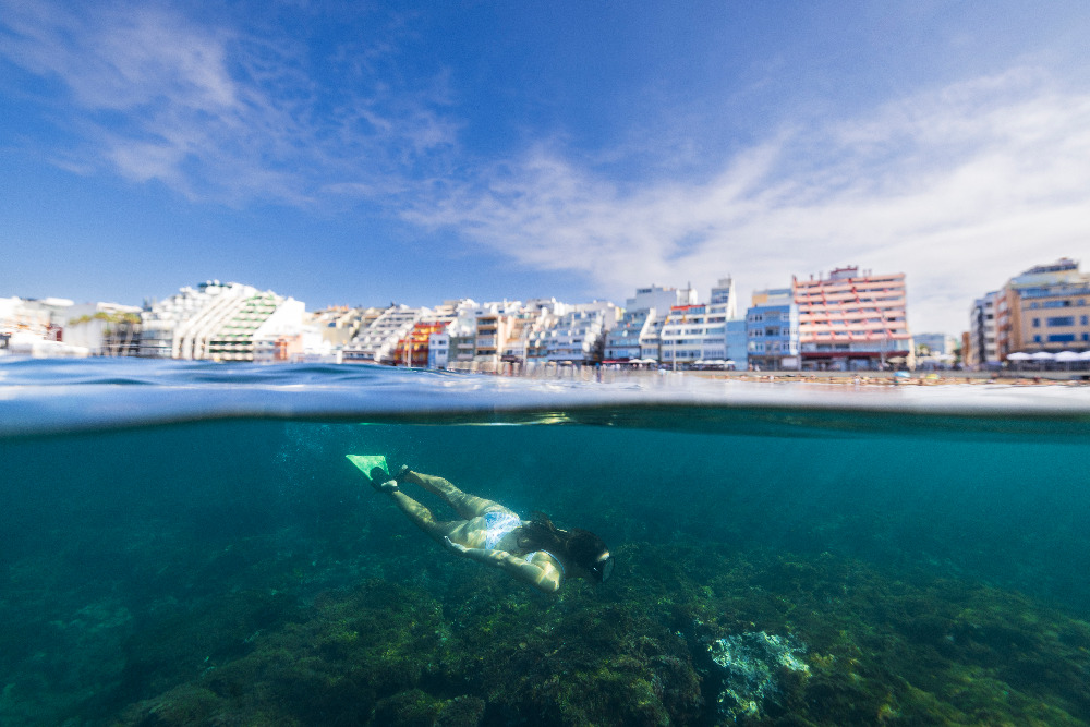 Schnorchlerin unter Wasser nahe der Küste, im Hintergrund Gebäude am Strand von Las Canteras.